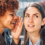 Eine Frau flüstert einer anderen etwas ins Ohr, beide lächeln - symbolische Darstellung für Lästern und den Austausch von Gerüchten.