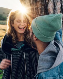 Ein lachendes Paar lehnt an einem Baum in der Sonne - Freundschaft und Gefühle verschwimmen.