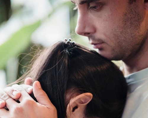 Eine Frau lehnt ihren Kopf an den Oberkörper eines Mannes, der traurig nach unten schaut und sein Kinn auf ihrem Kopf abstützt.