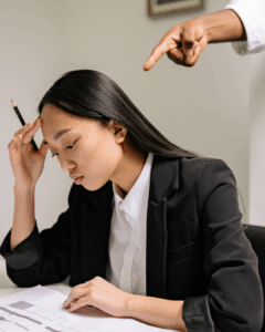 Foto einer Frau, die am Schreibtisch sitzt und einen traurigen und zweifelnden Gesichtausdruck hat. Hinter ihr ein Finger, der auf sie zeigt, als Symbolbild für den inneren Kritiker.