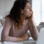 Foto einer braunhaarigen Frau, die vor dem Laptop sitzt und traurig und überfordert in die Ferne schaut. Sie spürt Versagensängste im Winter und einen Motivationsmangel.