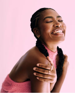 Frau in pinkem Oberteil die ein strahlendes Lächeln hat. Sie strahlt pure Selbstliebe und Zufriedenheit aus.