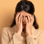 Frau mit braunen Haaren und beigem Pullover hält sich mit beiden Händen das Gesicht, wirkt traurig. Die Enttäuschung steht ihr ins Gesicht geschrieben. Der Hintergrund ist in warmem Gelb gehalten.