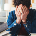 Überforderte Person im Büro mit Händen vor dem Gesicht, umgeben von Unterlagen und Laptop – Symbol für Angst vor Verantwortung, Stress im Job, Entscheidungsdruck und Überforderung im Alltag.