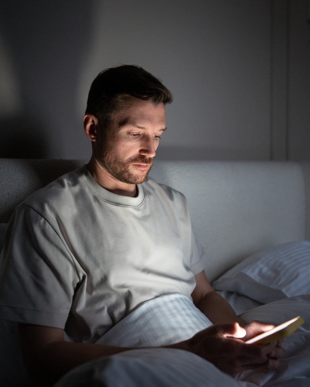 Mann, der im Bett sitzt und am Handy prokrastiniert. Er zeigt ein klares Bild von Selbstabotage.