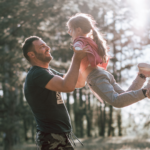 Nahaufnahme eines liebevollen Moments zwischen Vater und Tochter im Freien. Ein Vater hebt seine lachende Tochter spielerisch in die Luft, beide schauen sich lächelnd an. Warmes Sonnenlicht fällt durch die Bäume im Hintergrund und vermittelt Nähe, Vertrauen und emotionale Verbundenheit. Das Bild steht sinnbildlich für eine sichere, unterstützende Vater-Kind-Beziehung und emotionale Verfügbarkeit.