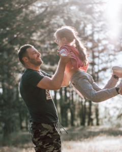 Nahaufnahme eines liebevollen Moments zwischen Vater und Tochter im Freien. Ein Vater hebt seine lachende Tochter spielerisch in die Luft, beide schauen sich lächelnd an. Warmes Sonnenlicht fällt durch die Bäume im Hintergrund und vermittelt Nähe, Vertrauen und emotionale Verbundenheit. Das Bild steht sinnbildlich für eine sichere, unterstützende Vater-Kind-Beziehung und emotionale Verfügbarkeit.