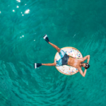 Warum Urlaub nicht immer erholsam ist: Psychologie der Regeneration 13 Person liegt entspannt auf einer Schwimminsel in türkisfarbenem Wasser und genießt eine ruhige Urlaubssituation aus der Vogelperspektive.