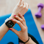Nahaufnahme einer Frau, die vor dem Sport ihre Smartwatch auf einer blauen Fitnessmatte überprüft. Der Fitness-Tracker zeigt Aktivitätsdaten und Trainingsziele an und steht symbolisch für digitales Gesundheitstracking, Selbstoptimierung und die Kontrolle der eigenen Bewegung.